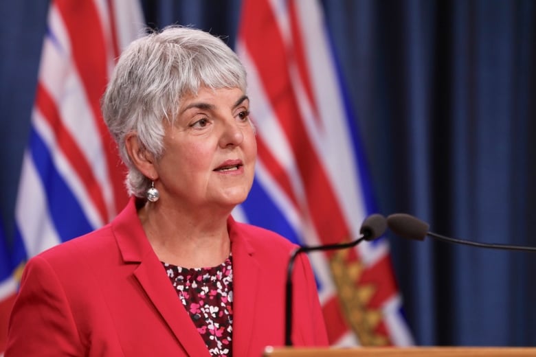 A woman with short white hair, wearing a red blazer speaks at a rostrum.