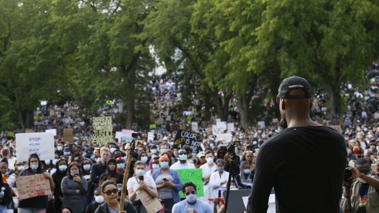 A Black man addresses a crowd of people, some holding protest signs. 