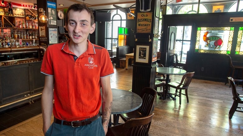 A man wearing a red shirt and grey pants stands inside a bar.