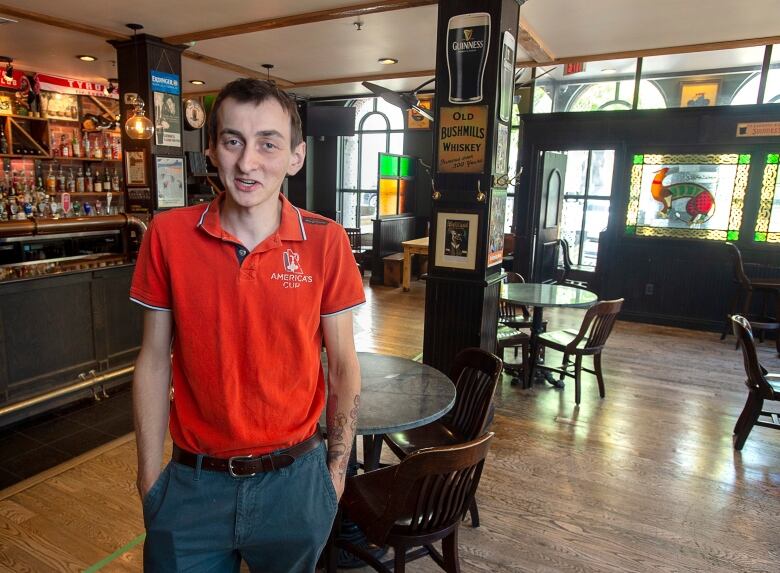 A man wearing a red shirt and grey pants stands inside a bar.