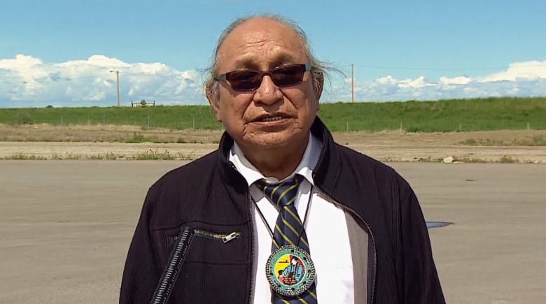 A man wearing an Indigenous pendant stands next to a country road.