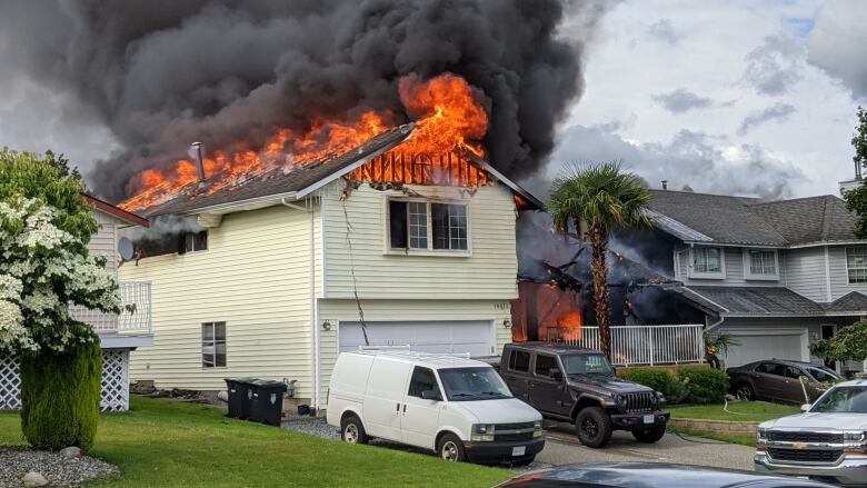 A yellow-sided single-family home with its roof in flames and a white van and a black truck parked in the front driveway. The house is next to a row of other similar homes.