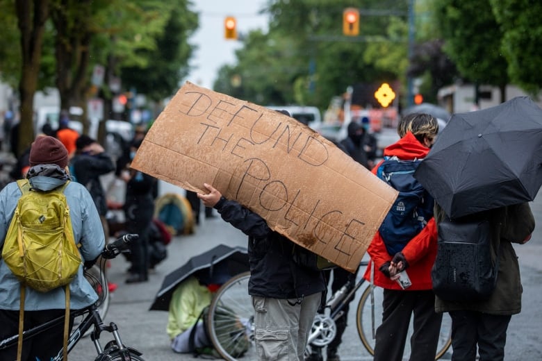 A person with a cardboard 'Defund the police' sign stands at the back of a procession during rainy weather.