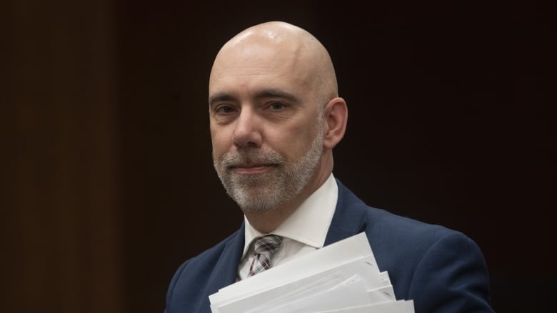 A bald man with a white grey beard who's wearing a blue suit holds a pile of papers.