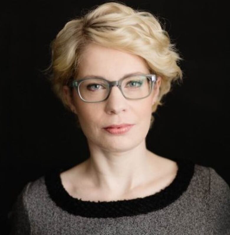 Woman with short blonde hair and glasses against black backdrop