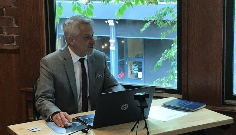 A man with short grey hair sits at a desk in front of a computer and is looking off in the distance to the right of the photo. He is wearing a grey suit, white dress shirt and dark tie.