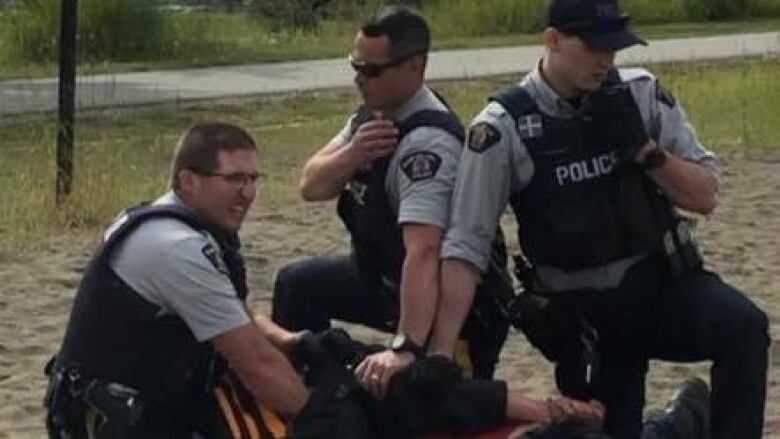 3 RCMP officers are seen holding a man to the ground on a beach volleyball court. 