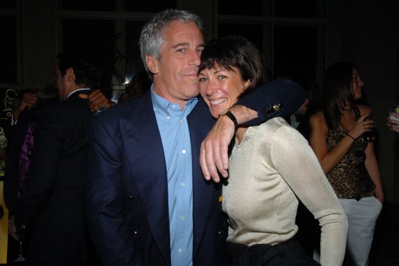 A white man with white hair stands with his arm around a woman with short black hair at night.