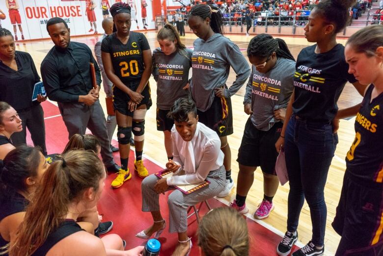 A woman in pants, a white blouse and heels sits in the centre of a huddle of female players on the edge of a basketball court, giving them instructions.