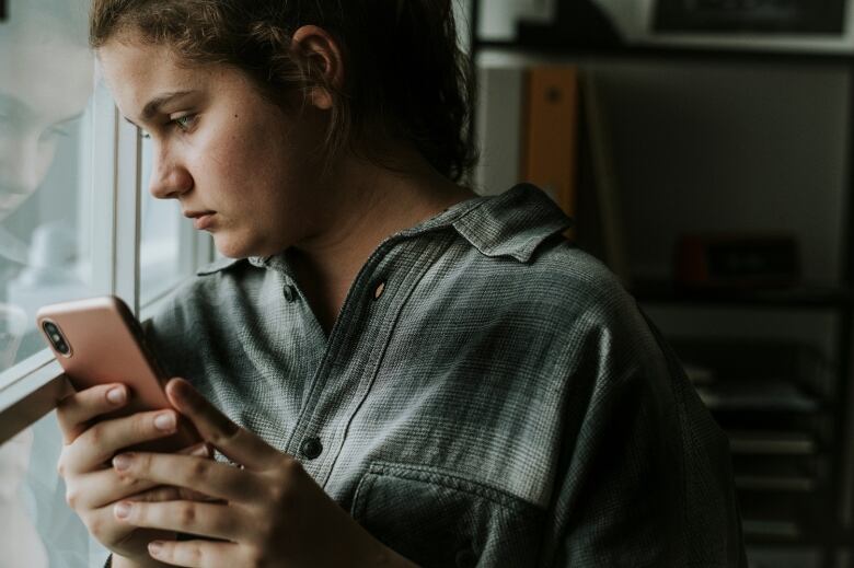 A young person holds a phone and looks out a window.