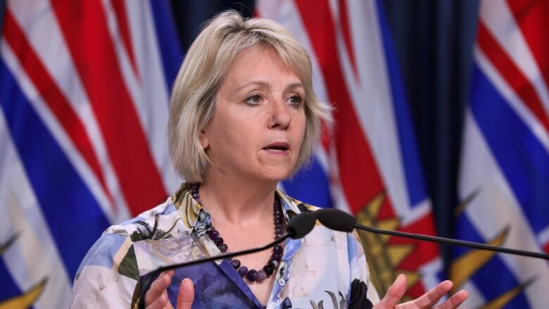 A white woman stands at a podium with the flags of British Columbia in the background.