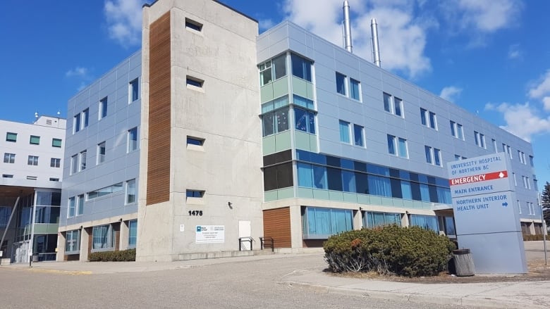 A blue and tan building with small windows and two narrow ventilation stacks on the roof. Beside it is a sign that says University Hospital of Northern B.C.