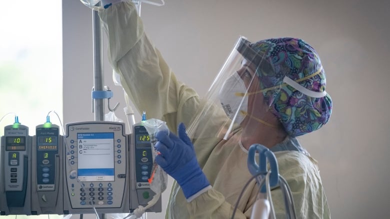 A nurse wearing full personal protective gear tends to a patient in an intensive care unit, checking the monitoring equipment and adjusting the IV bag.