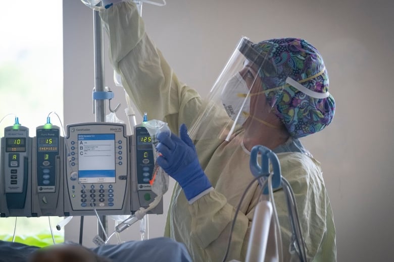 A nurse wearing full personal protective gear tends to a patient in an intensive care unit, checking the monitoring equipment and adjusting the IV bag.