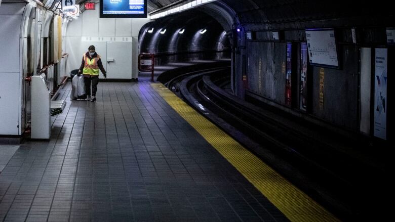 A transit employee wearing a yellow safety vest stands on an empty SkyTrain platform holding a garbage bag. 