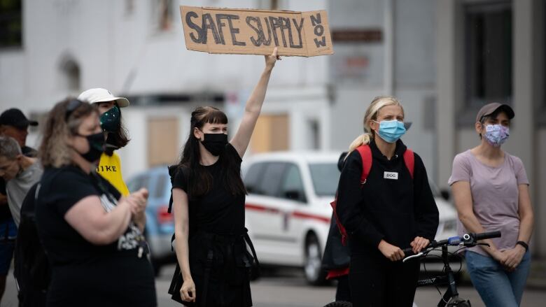 A person wearing a facemask holds up a sign that reads 'Safe Supply Now' at a rally.