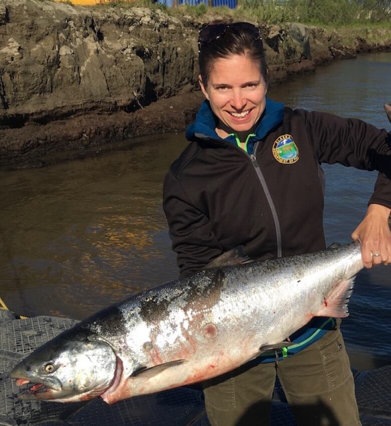 A woman in a jacket holds up a large fish by a river.