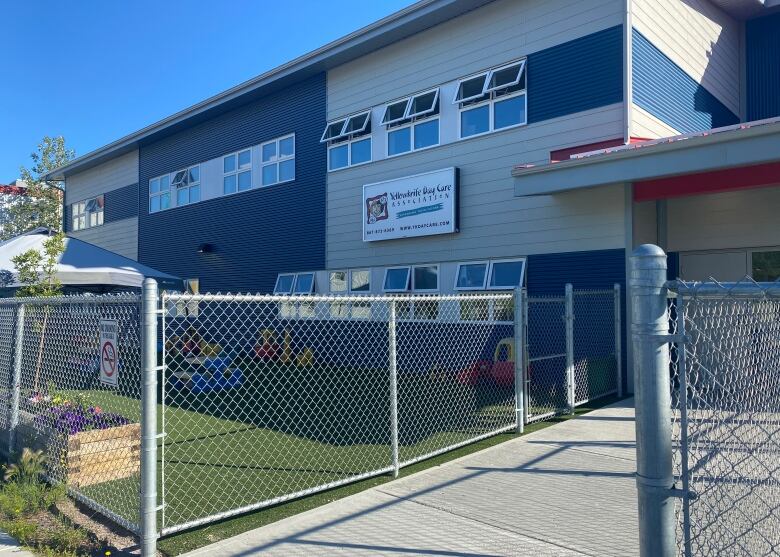 A blue and gray building stands with a wire fence around it and artificial grass on the ground. 