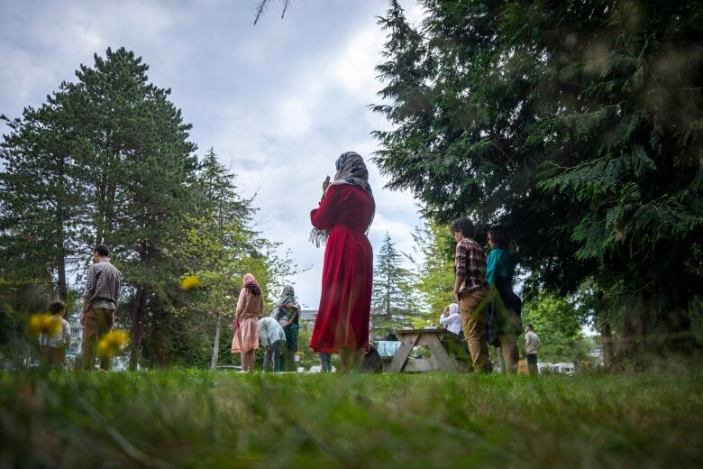 A small group of people prays in a park, with a woman in a red dress and grey hijab at centre.