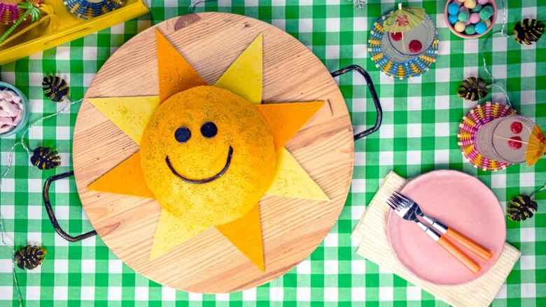 overhead shot of a round yellow cake decorated to look like a smiling sun. it's sitting on a green and white check tablecloth. 