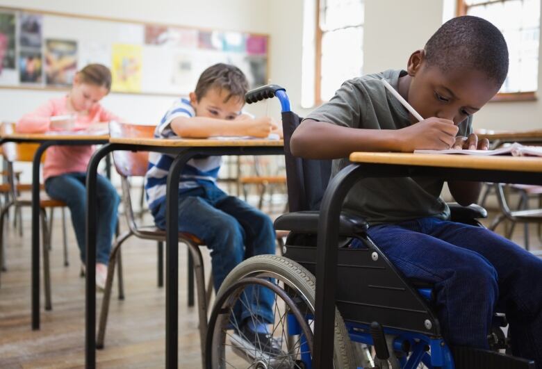 Three young students are shown writing at desk. The boy closest to the front is in a wheelchair.