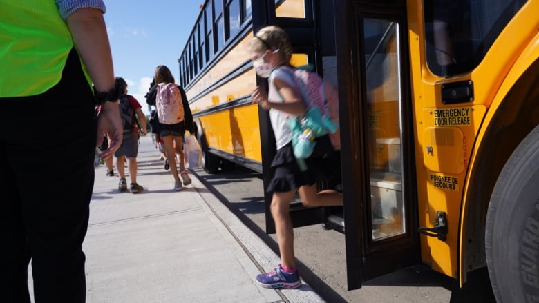 A child wearing a mask gets off of a school bus while several other children walk on the sidewalk.