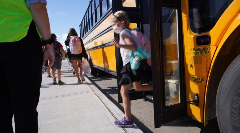 A child wearing a mask gets off of a school bus while several other children walk on the sidewalk.