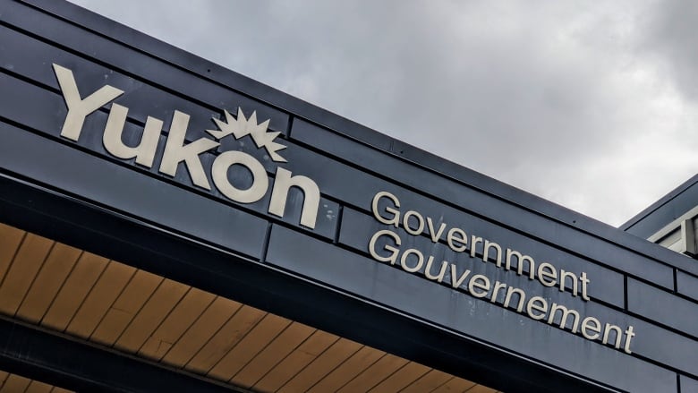 A building facade with the words Yukon government in the forefront of a grey sky.