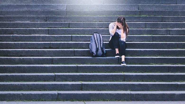 Teenager sitting on stairs with head in hands, backpack on ground next to her.