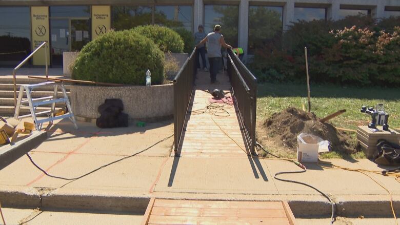 A person is shown walking towards the top of a wooden accessibility ramp with dark railings on both sides. The ramp is next to a set of concrete steps outside of a glass commercial building, the Elections NB returning office on Prospect Street.