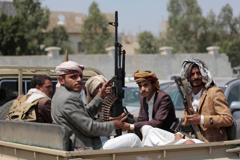 Five men, some armed with large machine guns, sit in the back of a pickup truck. 