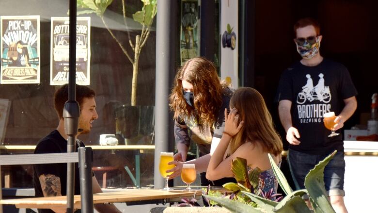 Servers deliver drinks to people sitting on a patio. Servers are wearing medical masks.