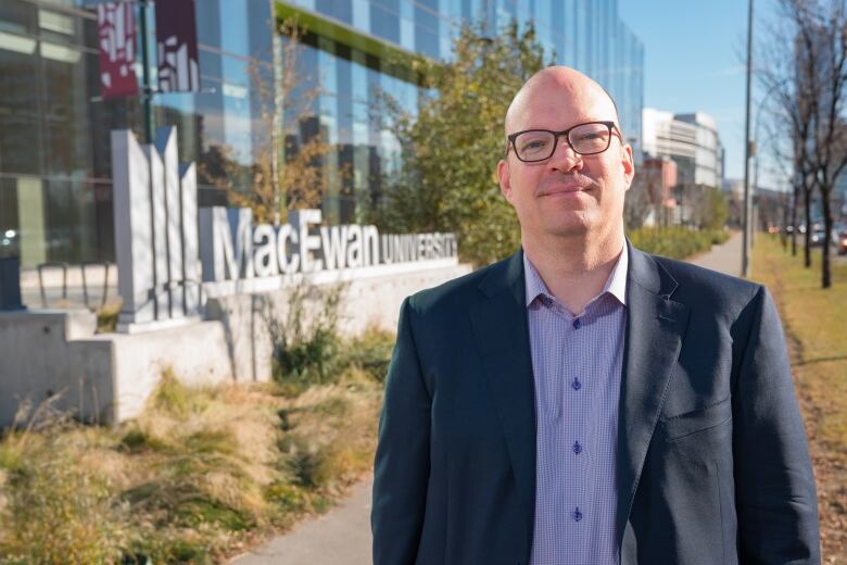 A man in a suit stands in front of a sign for MacEwan University.