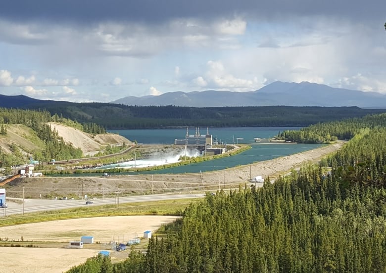 The headstock of the Whitehorse dam in front of Schwatka Lake. Water is rushing through the spillway. The scene is surrounded by boreal forest.