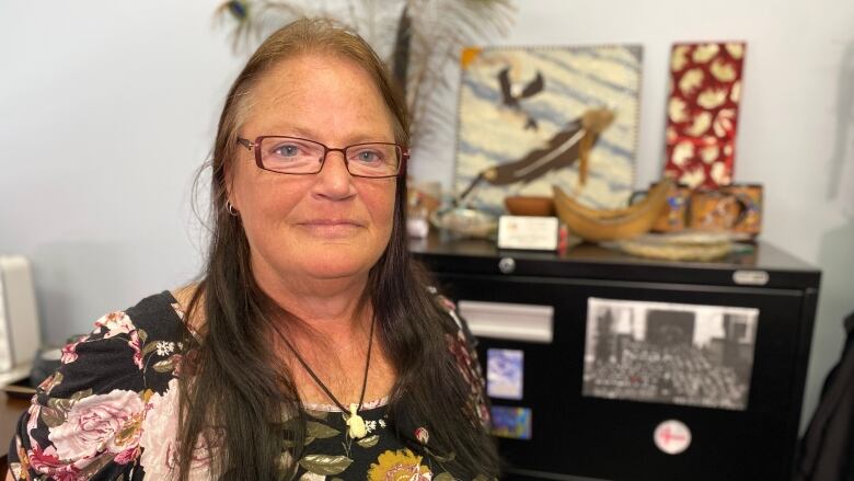 Woman in glasses and floral dress stands in front of a file cabinet on top of which are various pieces of Indigenous art. 