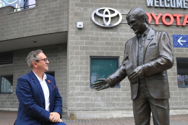 A man in a blue blazer glances, smiling, at a statue outside a stadium.