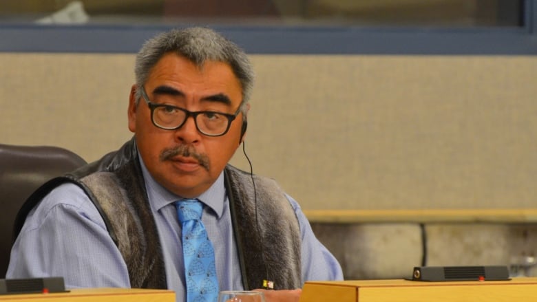 An Inuk man with salt and pepper hair sits in the Nunavut Legislature wearing a sealskin vest, matching his haircolour.