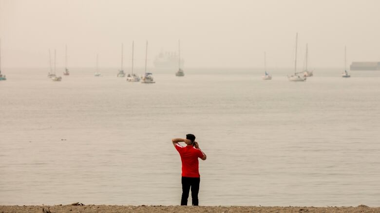 A man in  red shirt stands on a beach in Vancouver with his had on the back of his head while looking out to the ocean where pleasure boats are shrouded in wildfire smoke.