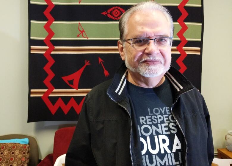 A man in a black zip-up sweater and dark shirt stands in front of a patterned blanket hanging on a wall.