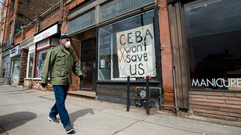 A person wearing a mask walks by storefronts, including one that displays a sign that says, 'CEBA won't save us.'