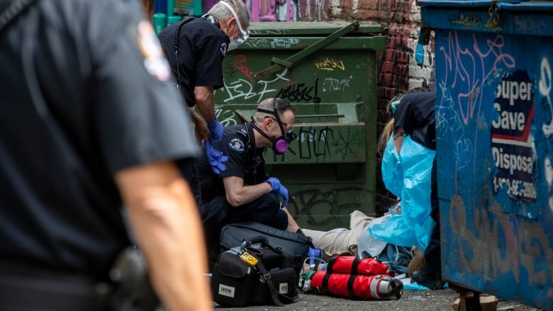 Paramedics attend a person suspected of having a drug overdose beside a dumpster in an alley.