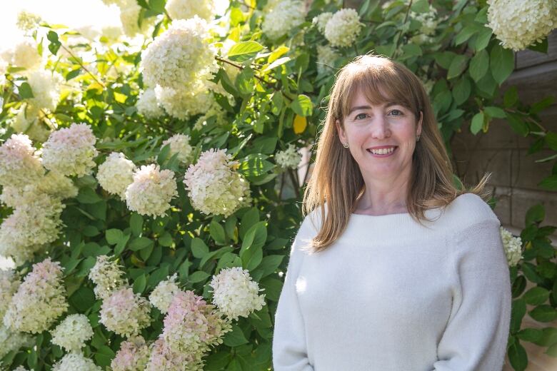 A woman in a white sweater with long blonde hair smiles for the camera in front of a flower bush with large white blossoms.