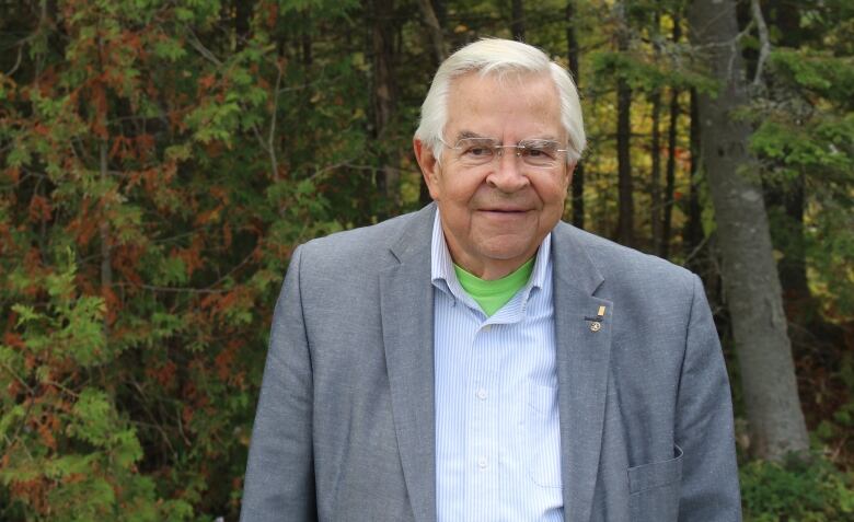 A man in a grey suit smiles at the camera outside with trees behind him.