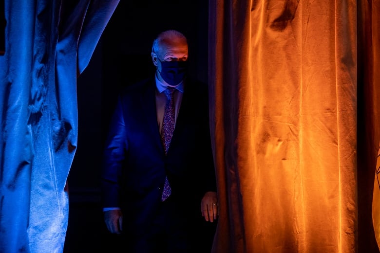 A man in a mask comes out from behind a curtain on a lit stage.