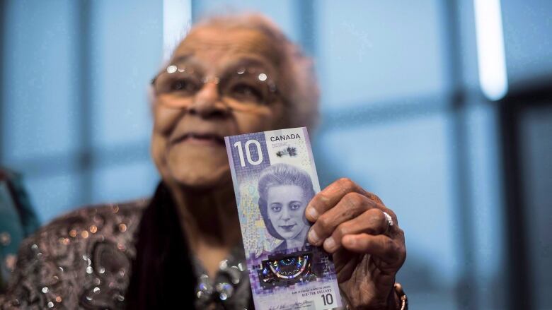 An older Black woman holds a 10 dollar bill with the face of Viola Desmond on it