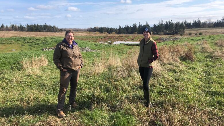 Two women stand in a field with a wetland behind them.