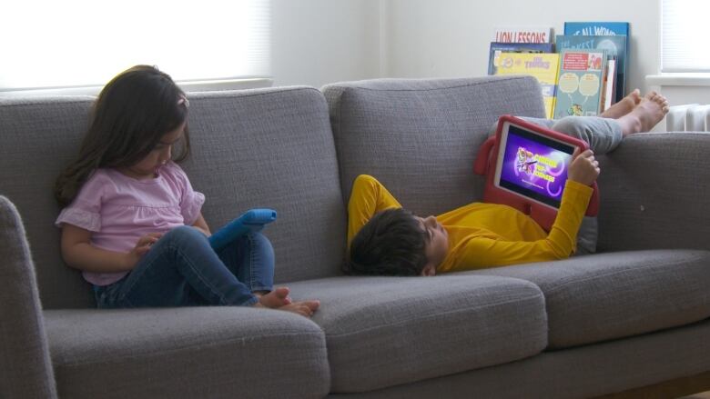 Two children sit on a couch, looking at tablets