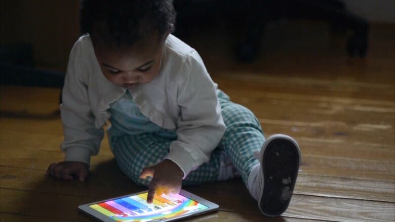 A toddler sits on the ground pointed at a tablet with a colourful screen