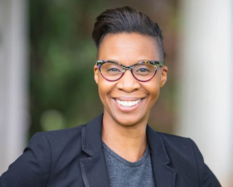 A person wearing a dark-coloured blazer stands outside and smiles.