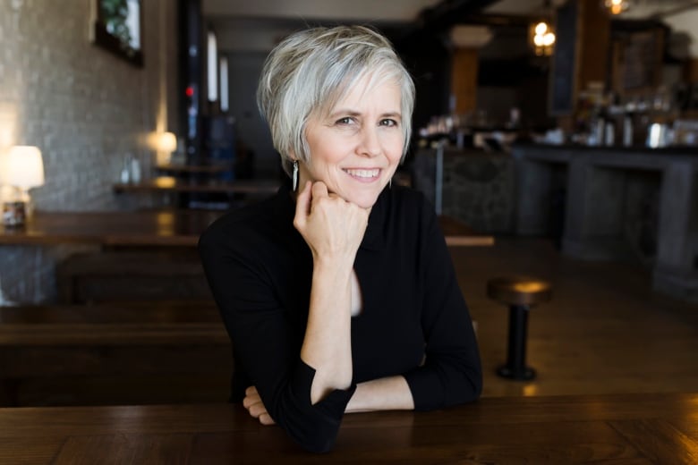 Shauna Powers sits in a restaurant with low lighting, dark wooden tables and white stone walls. She has white hair and is wearing a black cardigan.